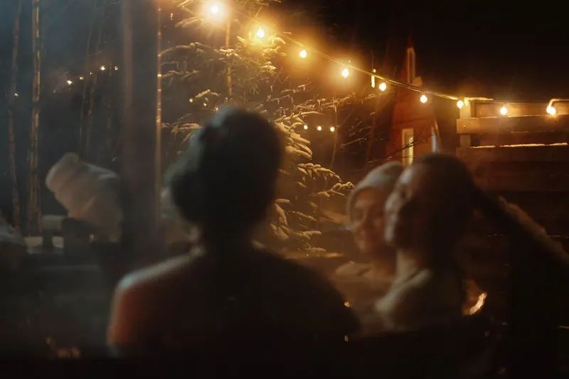 Three people enjoy a warm and cozy night in an outdoor hot tub surrounded by snow and string lights